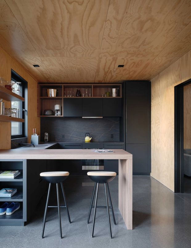 Compact contemporary kitchen with a peninsula breakfast bar and two stools. The space features extensive natural plywood wall and ceiling lining, dark matte cabinetry, open shelving, a dark stone-look splashback, integrated appliances, and a polished grey concrete floor. A doorway at right leads toward an adjacent room.