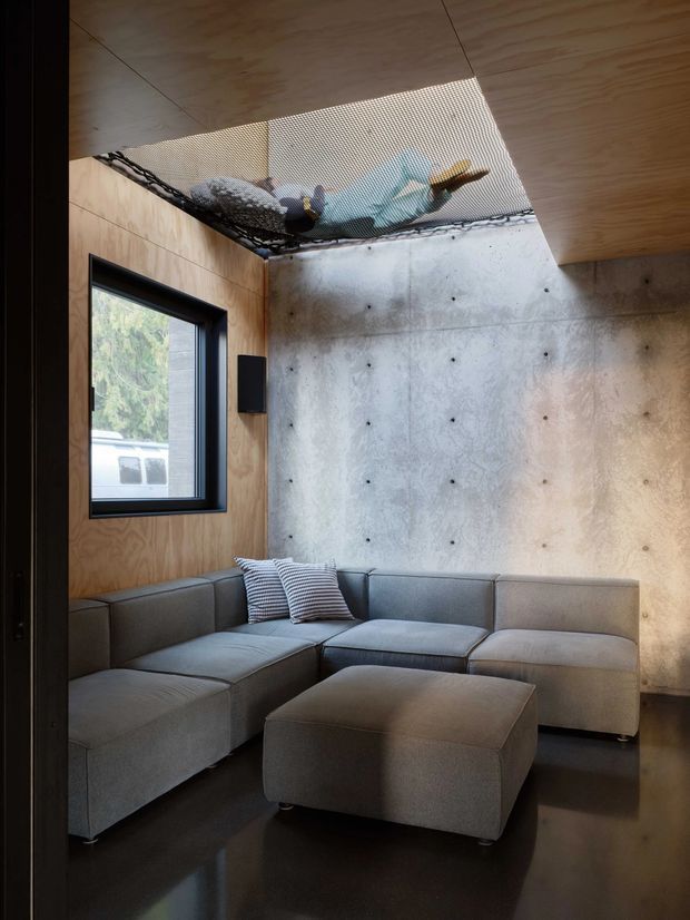 A compact, modern lounge with a grey sectional sofa and matching ottoman arranged against a raw concrete feature wall. Plywood wall and ceiling panels, dark polished flooring, a black-framed window, and an exposed mesh ceiling detail create an industrial, contemporary interior.