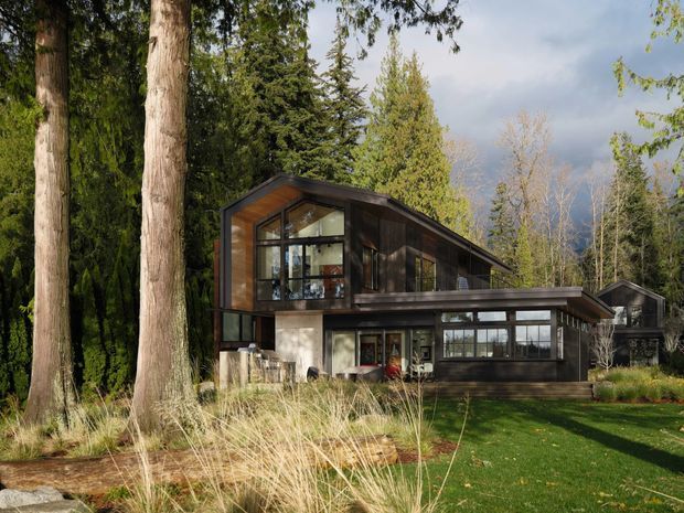 A contemporary multi-level house set within a wooded landscape. The dark timber-clad upper volume has a steep asymmetrical roof and extensive black-framed glazing, while the lower level combines dark cladding, concrete walls, and a broad projecting roofline. Tall mature trees, ornamental grasses, and a lawn surround the home.