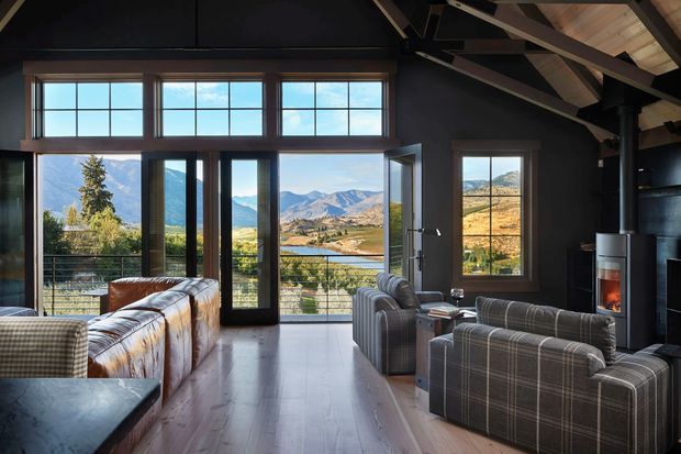 A dramatic double-height living room with expansive floor-to-ceiling glazing and balcony access overlooking a mountainous valley. The room features dark painted walls, exposed timber roof structure, pale timber flooring, leather and plaid upholstered sofas, and a freestanding wood-burning stove. Large windows frame the surrounding alpine landscape.