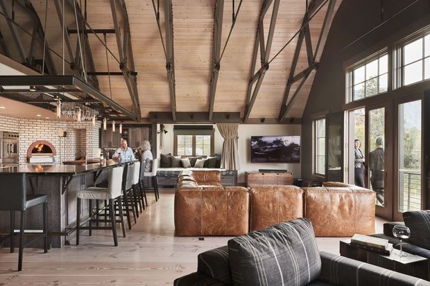 Open-plan barn-style living space with a kitchen and island bar on the left, a lounge area with large brown leather sofas in the centre, and full-height glazed doors and windows on the right. The room has exposed dark timber/steel roof trusses, a pale timber-lined pitched ceiling, wide timber flooring, dark-framed glazing, and a white brick kitchen wall with a built-in pizza oven. The space is warm, spacious, and rustic-industrial in character.