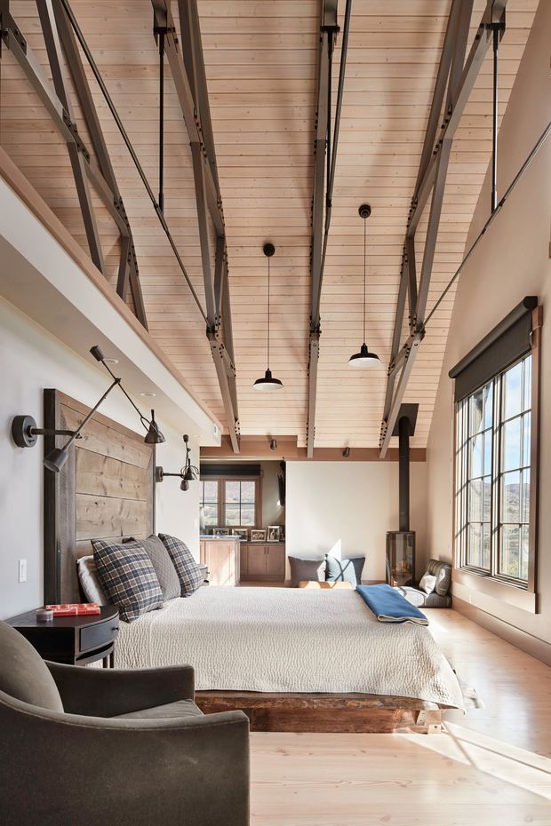 A spacious, warmly lit bedroom with a high exposed timber ceiling and prominent dark metal structural beams. The room features a large bed with a textured light bedspread, a rustic reclaimed timber headboard, plaid cushions, dark upholstered armchairs, wall-mounted reading lights, pendant lighting, wide industrial-style windows, and a small kitchenette or sitting area visible at the rear. The design blends rustic materials with industrial detailing and contemporary furnishings.