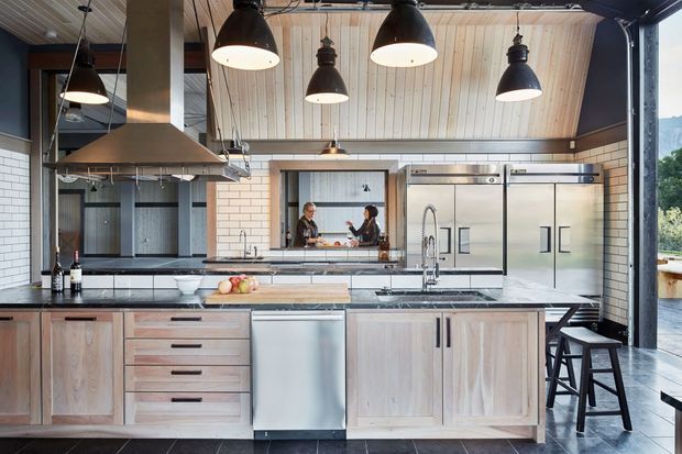 A spacious industrial-style kitchen with light timber cabinetry, dark stone benchtops, stainless-steel appliances, and a large central island. The room features a high pitched timber-lined ceiling, exposed services, multiple black pendant lights, a substantial stainless-steel rangehood, and white brick walls. Large openings provide views to the surrounding landscape and an adjacent outdoor area.