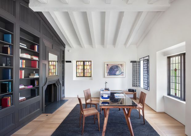Bright, refined dining room with a large dark built-in bookcase and fireplace surround along the left wall, a timber dining table with upholstered wooden chairs, and a dark area rug over a light timber floor. White painted exposed ceiling beams, white walls, decorative leaded or stained-glass windows, and framed artwork create a traditional architectural character with contemporary furnishings.