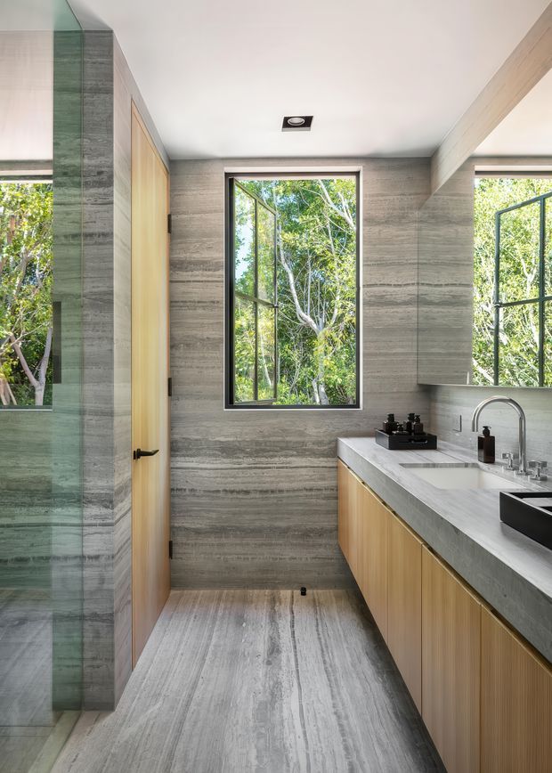 A compact, refined bathroom with a walk-in glass shower on the left and a long timber-veneer vanity on the right. The vanity has a pale grey stone countertop, integrated rectangular basin, and brushed metal gooseneck tapware. Grey horizontally veined stone or porcelain surfaces continue across the walls and floor, while a natural timber door and black-framed opening windows provide contrast. Lush trees are visible through the glazing, creating a strong connection to the outdoors.