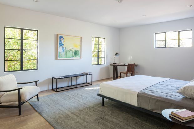 Bright, spacious bedroom with white walls, black-framed grid windows, a low dark bed, a large neutral rug, a minimalist console, a small writing desk, and a sculptural lounge chair. Natural light and restrained furnishings create a clean, calm modern interior.