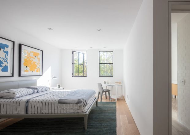 Bright, minimalist bedroom with a low-profile upholstered bed dressed in blue-grey striped bedding, framed botanical artwork, pale timber flooring, and a dark textured area rug. Two tall black-framed windows provide natural light, while a compact white desk and chair sit at the far end. An open doorway reveals a partially visible adjacent space with white brickwork and timber cabinetry.