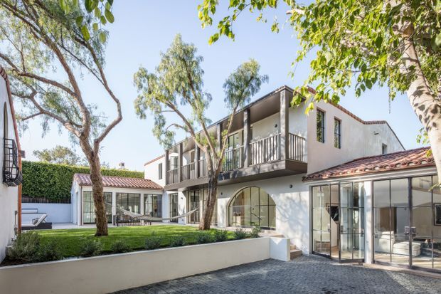 A sunlit multi-level residential courtyard viewed from outside, with white rendered walls, dark-framed glazing, red terracotta-tiled roofs, upper-level balconies, and mature trees surrounding a paved forecourt. The architecture combines contemporary additions with Mediterranean-inspired roof forms and a landscaped courtyard setting.
