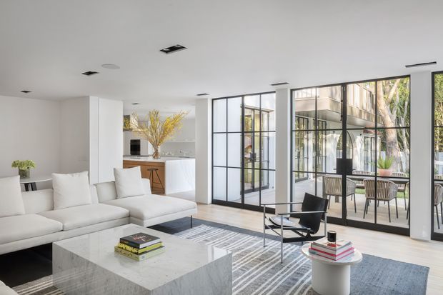 Bright open-plan living room with a large white sectional sofa, sculptural marble coffee table, patterned area rug, and a black lounge chair. Black-framed glazed partitions and doors open onto a furnished outdoor terrace, while a compact white kitchen with a timber breakfast stool is visible in the background. The space has a clean, restrained contemporary fit-out with abundant natural light.