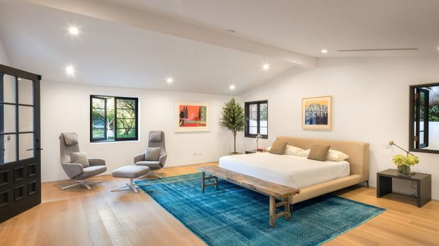 Spacious, bright bedroom with a low-profile upholstered bed, layered neutral bedding, timber flooring, and a large blue area rug. The room includes two lounge chairs with ottomans, a timber coffee table/bench, a dark bedside table, potted greenery, framed artwork, black-framed windows, and a glazed black interior door. Recessed ceiling lighting and a restrained neutral palette create a contemporary, refined atmosphere.