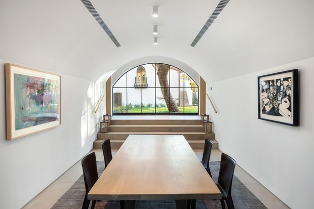 Bright contemporary dining room with a long light-wood dining table and dark upholstered chairs. White walls and ceiling frame a distinctive arched, black-framed window overlooking greenery, with stepped seating or a low platform beneath it. Recessed ceiling lighting, linear air vents, framed artwork, and a neutral rug complete the refined space.