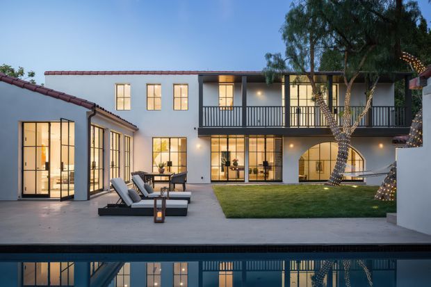 Twilight view of a refined two-storey courtyard residence organised around a rectangular lawn and reflecting pool. The white rendered facade has dark-framed floor-to-ceiling glazing, a covered balcony with dark vertical balustrades, warm interior lighting, and a low-pitched tiled roof over the single-storey wings. Outdoor loungers and subtle tree lighting create an inviting, resort-like atmosphere.