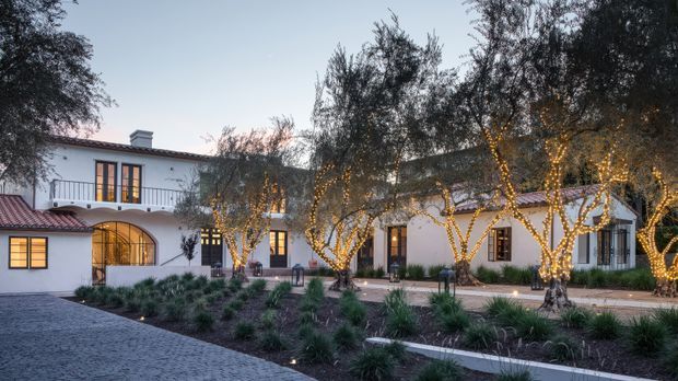 Twilight exterior view of a large white stucco residence arranged around a landscaped courtyard. The building has red clay tile roofing, arched openings, dark-framed windows and doors, and a balcony railing. Olive trees wrapped in warm string lights line the paved courtyard, with ornamental grasses and low planting beds in the foreground.
