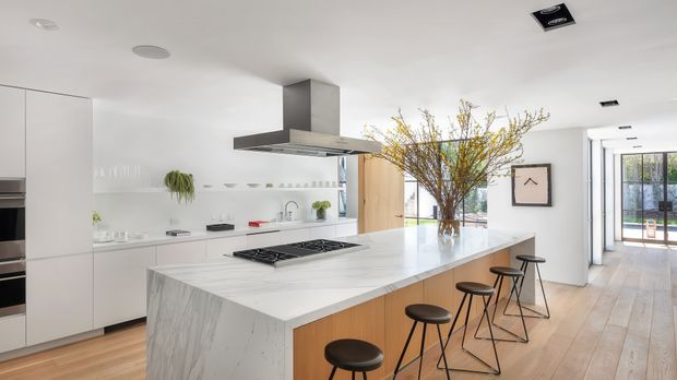 Bright, spacious contemporary kitchen with a large white marble-look island, integrated white cabinetry, a central stainless-steel rangehood, and black bar stools. The room opens toward a glazed hallway or outdoor area, with pale timber flooring, minimalist detailing, and abundant natural light.