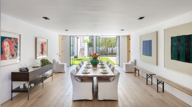 Bright contemporary open-plan dining and living space with a long timber dining table, upholstered dining chairs, pale timber flooring, white walls, large abstract artwork, streamlined console furniture, and full-height glazed doors opening to a landscaped courtyard. The room has a clean, gallery-like appearance with abundant natural light.