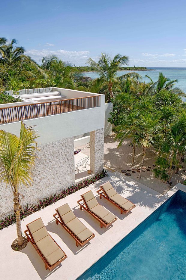 Elevated exterior view of a contemporary coastal resort-style property with a rooftop terrace, pale textured masonry walls, timber-toned vertical railings, tropical palm landscaping, a pool deck with multiple sun loungers, and a swimming pool overlooking turquoise ocean water.