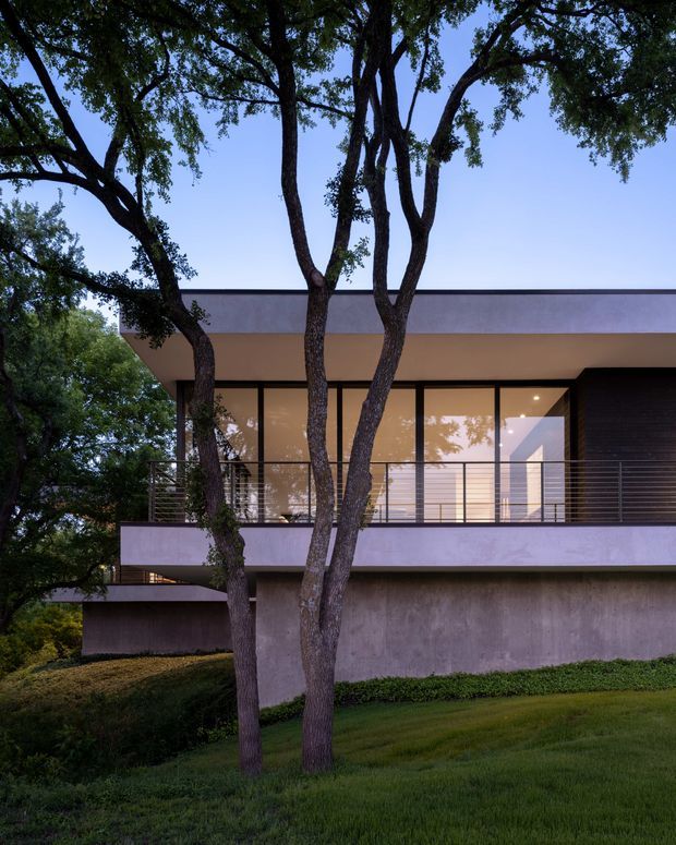 A modern two-storey residential facade set among mature trees and landscaped lawn. The building features broad horizontal cantilevered slabs, a flat roof, extensive floor-to-ceiling glazing, slender dark window frames, a metal-railed balcony, and smooth light grey and white rendered surfaces. Warm interior lighting is visible through the glazed upper level.