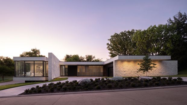 Single-storey contemporary residence viewed from the street at dusk. The building has a low, predominantly flat roofline, crisp geometric volumes, extensive floor-to-ceiling glazing in the left wing, pale textured masonry walls, dark recessed openings, and a timber-look vertical screen at the central entry or courtyard zone. A landscaped planted strip and paved driveway occupy the foreground.