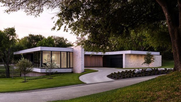 A low-profile modern single-storey residence set within a landscaped suburban site. The home has crisp flat rooflines, pale masonry walls, expansive floor-to-ceiling glazing, a recessed dark garage, and a curved concrete driveway bordered by lawn, planting, and mature trees. The image is an exterior architectural photograph taken at dusk.