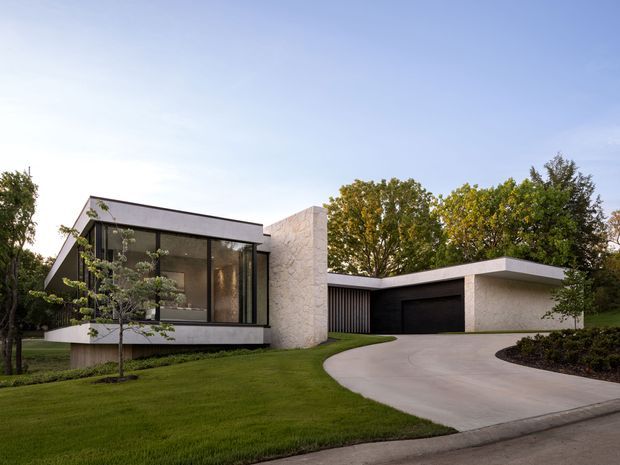 Exterior view of a low-slung modern house with a flat-roofed composition, expansive dark-framed glazing, pale textured masonry walls, and an attached single-car garage. A curved concrete driveway leads through a carefully landscaped lawn and garden, with mature trees surrounding the property.