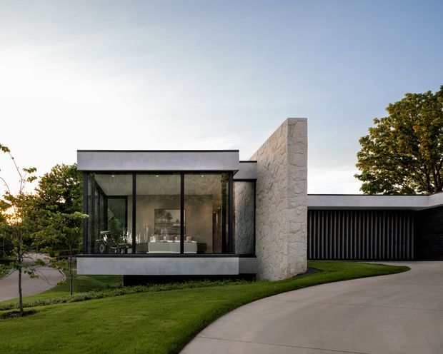 Exterior view of a contemporary single-storey residence set in landscaped grounds. The design features a flat roof, extensive floor-to-ceiling glazing in a projecting living volume, a tall light stone-clad wall, dark vertical slatted screening, and a curved concrete driveway.