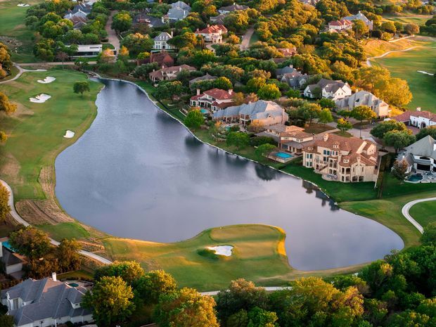 Aerial photograph of an upscale residential neighbourhood arranged around a large lake or water feature, with expansive manicured golf-course fairways, sand bunkers, curving roads, mature trees, and numerous detached homes. The scene is photographed in warm late-day light and presents a landscaped suburban estate setting.