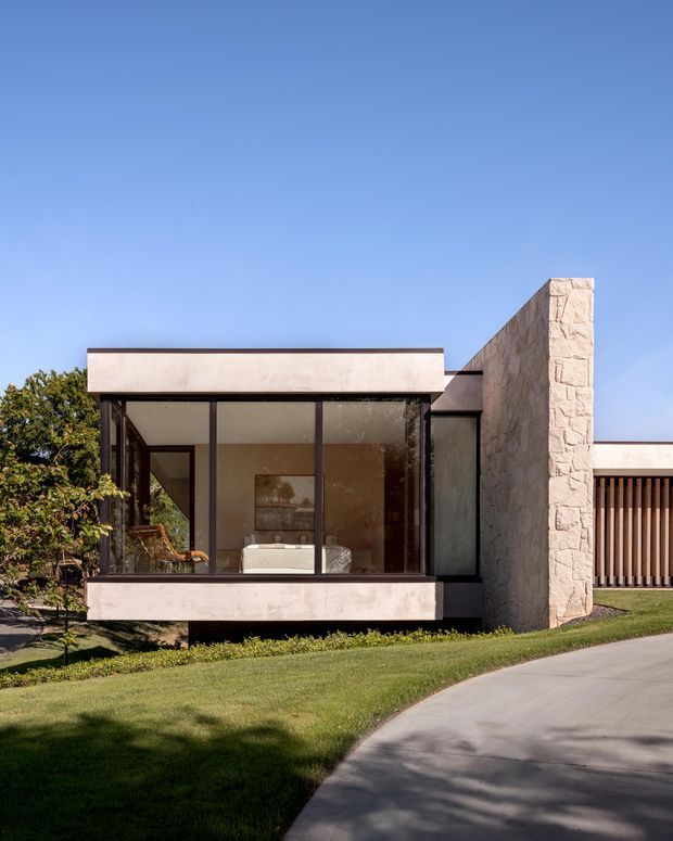 Exterior view of a low, modernist house with a flat-roofed glazed living space, dark-framed floor-to-ceiling windows, pale rendered horizontal bands, and a prominent light stone-clad vertical wall. A timber slatted screen and driveway are visible to the right, with landscaped lawn and mature planting around the building.