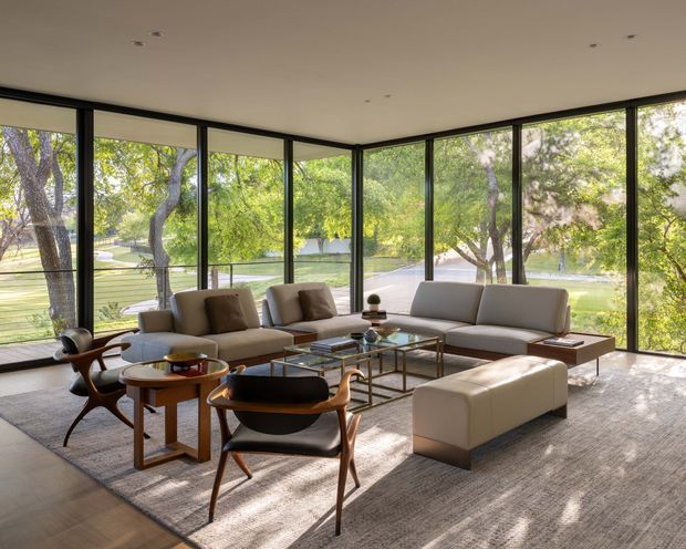 A bright, spacious living room with floor-to-ceiling black-framed glazing on two sides, overlooking a leafy landscaped setting. The room features low-profile contemporary sofas, sculptural timber-and-leather lounge chairs, nested side tables, a glass coffee table, a large upholstered ottoman, and a neutral textured rug. Recessed ceiling lighting and minimal detailing create a refined mid-century-inspired contemporary interior.
