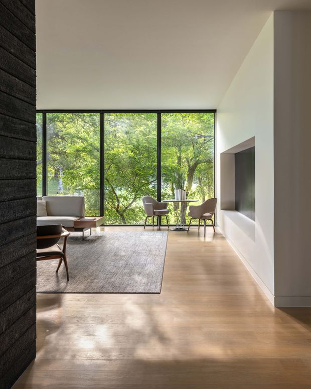 A refined contemporary living room with full-height black-framed glazing overlooking leafy trees. The room features warm timber flooring, white walls, a large neutral area rug, a light sofa, sculptural lounge chairs, a small dining or occasional table, and a recessed dark feature wall or television niche. A dark textured partition wall frames the left side of the image. Natural light creates a calm, airy interior.