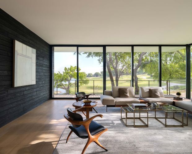 A refined modern living room with expansive black-framed floor-to-ceiling glazing overlooking a leafy landscaped setting. The room features a dark horizontal timber feature wall, warm timber flooring, a neutral sectional sofa, sculptural mid-century-style lounge chairs, round wooden tables, a glass coffee table with a metal frame, and a large pale area rug. The space is bright, open, and visually connected to the outdoors.