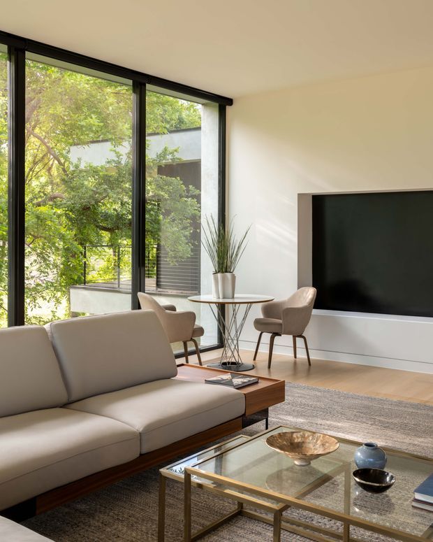 A bright, refined contemporary living room with floor-to-ceiling black-framed glazing opening toward leafy greenery. The room features a streamlined cream sectional sofa with a timber base, two upholstered lounge chairs around a small round table, a large wall-mounted black television, a glass-topped coffee table with a metal frame, pale timber flooring, and layered neutral rugs. The space has a clean, calm, indoor-outdoor feel with abundant natural light.