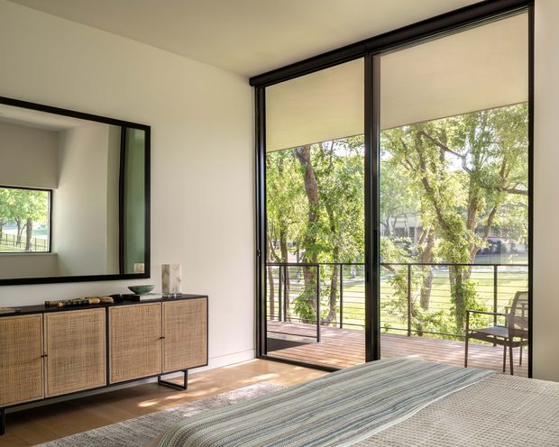 Bright contemporary bedroom with a low woven-front storage console, large black-framed glazed doors, roller blinds, and direct access to a timber balcony overlooking leafy grounds. A bed occupies the foreground, while a large mirror reflects the room and window.
