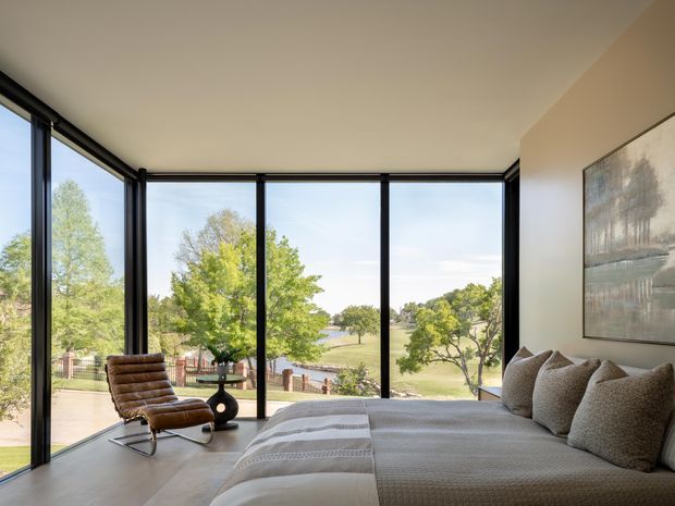 A serene contemporary bedroom with floor-to-ceiling black-framed glazing overlooking a leafy landscape and water. The room features a large upholstered bed with neutral textured bedding, a brown leather lounge chair, pale flooring, warm off-white walls, and abstract wall art.