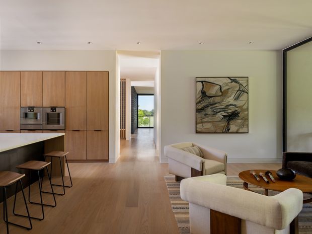 Open-plan contemporary kitchen and living area with warm timber cabinetry and engineered-looking timber flooring. The kitchen includes integrated stainless-steel appliances, a dark benchtop, and a peninsula with bar stools. The adjoining living space has curved upholstered armchairs, a round timber coffee table, a patterned rug, abstract artwork, and a large dark-framed glazed opening. A hallway extends toward a bright window beyond.