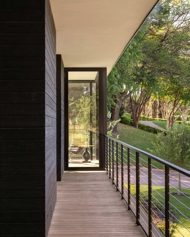 A covered elevated exterior walkway or balcony with dark horizontal-clad walls, timber decking, a slim dark metal balustrade, and a large frameless glazed opening at the end. The walkway overlooks a landscaped garden with mature trees and a curving path. The architecture is clean-lined and minimalist, with a strong indoor-outdoor connection and warm natural light.