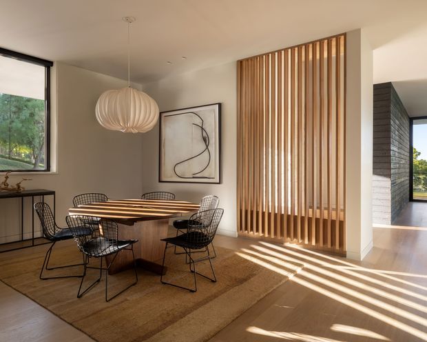 Warm contemporary dining area with a round timber dining table and wire-frame chairs on a natural woven rug. A sculptural pendant light hangs above, while a full-height timber slatted screen filters strong sunlight into the room. Neutral plaster walls, timber flooring, minimalist artwork, and a glimpse toward an adjacent space create an airy, refined interior.