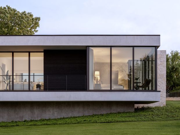 Exterior view of a contemporary single-storey residence with a flat roof, strong horizontal lines, extensive floor-to-ceiling glazing, a projecting upper volume, dark feature cladding, and a pale stone-clad side wall. The house is set above a landscaped lawn, with a balcony or terrace visible through the glazed facade and interior furnishings visible behind the windows.