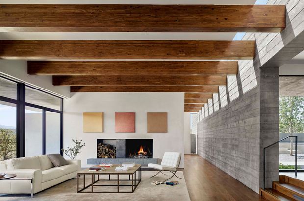 A spacious contemporary living room with exposed dark timber ceiling beams, a long textured concrete feature wall, floor-to-ceiling glazing, timber flooring, and a central fireplace. The room contains a light sectional sofa, coffee table, lounge chair, abstract wall art, and a stair opening at the right, creating a warm yet minimalist indoor-outdoor atmosphere.