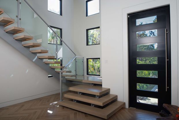 Bright contemporary entryway featuring a floating timber staircase with glass balustrades and slim metal supports, a dramatic black front door with narrow horizontal glazing panels, high-set windows, and a herringbone timber floor. The stair geometry and abundant glazing create an airy, refined double-height feel.