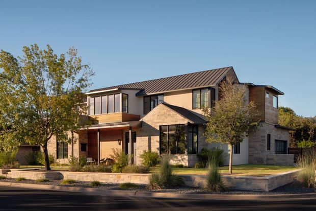 A two-storey contemporary residential house viewed from the street, with a long pitched metal roof, projecting upper-level volumes, extensive dark-framed glazing, and a mix of warm brick and pale masonry cladding. The landscaped frontage includes ornamental grasses, mature trees, low retaining walls, and a paved entry path.