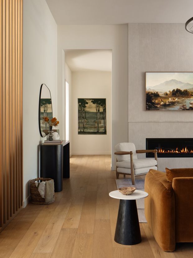Warm, refined living room with a contemporary fireplace wall, pale vertical wall panelling, wide light timber flooring, and a sculptural black console with mirror in the entry area. The room includes a caramel-brown sofa, upholstered lounge chair, small round side table, artwork, and soft layered lighting. A hallway or passage extends beyond the living area.