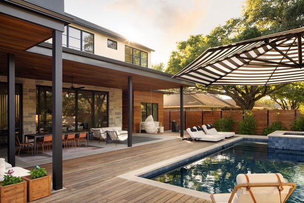 Contemporary two-storey residence with a covered outdoor living area opening onto a timber deck and rectangular swimming pool. The home features extensive glazing, warm timber soffits and decking, exposed brick walls, dark slender structural columns, and a large striped retractable shade canopy. Outdoor dining and lounge furniture frame the pool terrace, with mature trees and fencing creating a private landscaped setting.