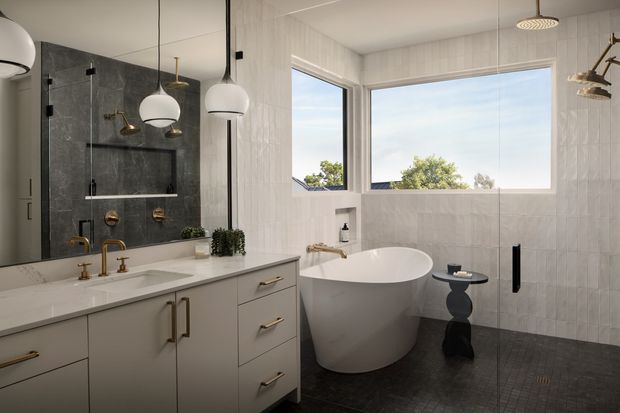 A bright, refined contemporary bathroom featuring a freestanding oval bathtub beside large windows, a wall-mounted shower, double vanity with brass hardware and fixtures, mirrored cabinetry, and dark textured floor tiles. White vertically textured wall tiles and warm brass accents create a polished, high-contrast scheme.