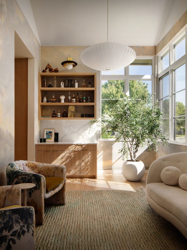 A sunlit, compact living room with large wraparound windows, warm textured plaster walls, light timber flooring, and a built-in timber bar or display cabinet with open shelving. The room features a sculptural white pendant light, a patterned floral armchair, a rounded cream sofa, a large potted tree, and a textured neutral rug. The overall atmosphere is warm, refined, and eclectic, with strong natural light and garden views.
