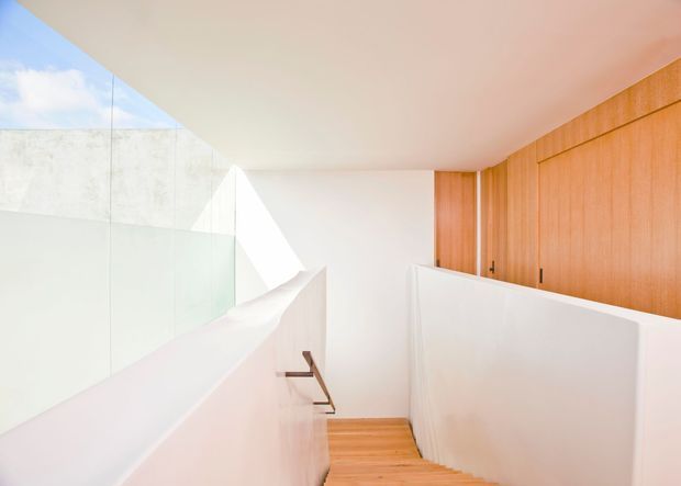 Bright minimalist interior stairway with warm timber flooring and a full-height timber-panelled wall. White walls and balustrades create a clean, sculptural composition, while a glass balustrade and skylight or clerestory glazing bring in abundant natural light. A simple dark metal handrail runs down the stairs.