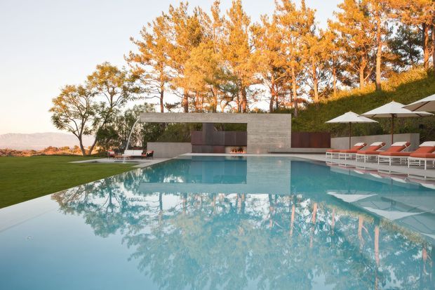A luxurious outdoor swimming pool dominates the foreground, reflecting autumn-coloured trees and a minimalist concrete pool pavilion. White sun umbrellas and red-orange loungers line the pool edge, with manicured lawn, clipped hedging and distant low hills surrounding the property. The architecture features clean horizontal lines, a flat roof and a recessed open entertaining area with a linear fire feature.