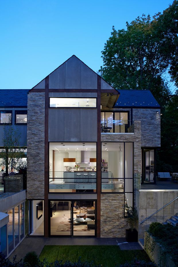 Twilight exterior view of a contemporary multi-storey residence with a prominent steep gable-front form, extensive floor-to-ceiling glazing, dark metal framing, and stacked-stone cladding. The illuminated interior reveals a kitchen and living areas, while terraces, stairs, retaining walls, and landscaped garden areas surround the house.