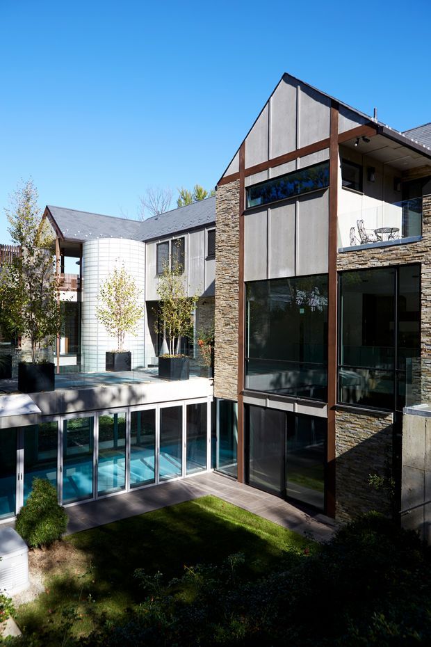 Exterior photograph of a multi-level contemporary residence arranged around a landscaped courtyard and swimming pool. The building combines dark vertical panel cladding, extensive black-framed glazing, stacked-stone wall sections, exposed dark timber or steel framing, and pitched roof forms. A covered balcony and terrace are visible, with a bright blue sky and mature planting surrounding the home.