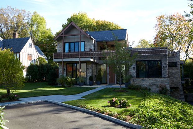 Exterior view of a multi-level contemporary house set within a landscaped suburban garden. The dark gabled main volume combines stone or masonry walls, extensive glazing, and a timber-toned balcony railing, while a lower side wing has a broad flat roof terrace and large dark-framed windows. A paved driveway and walkway lead to the home, with a neighbouring gabled house partially visible at left.