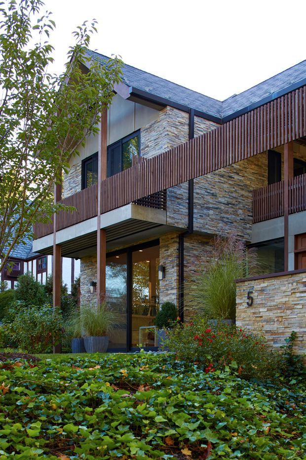 Exterior view of a contemporary multi-storey residence with a stepped, angular roofline, extensive stacked-stone wall cladding, dark metal-framed glazing, timber vertical privacy screens and balconies, and landscaped planting in the foreground. A visible house number is mounted on the stone boundary wall.